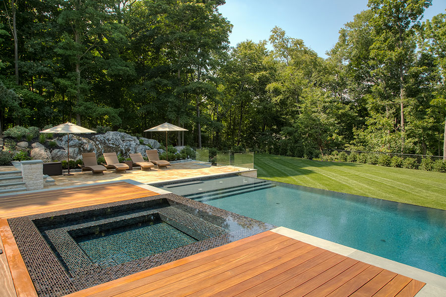 Elegant and minimalistic infinity pool featuring a wooden deck and jacuzzi, adorned with pool umbrellas and chairs, showcasing the exquisite pool renovation services in NY, NJ, and CT.