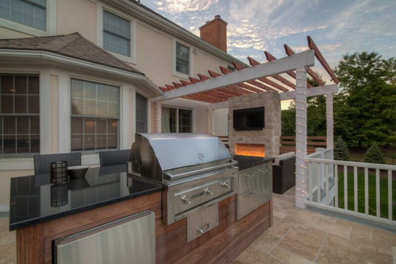 A modern outdoor kitchen with a stainless steel grill, granite countertops, and a pergola featuring string lights and a fireplace