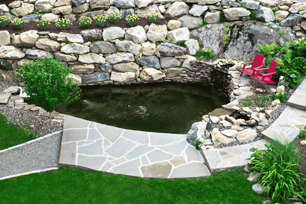 Naturalistic pond-like water feature with stone landscaping, adjacent to a patio and red chairs in a lush backyard setting