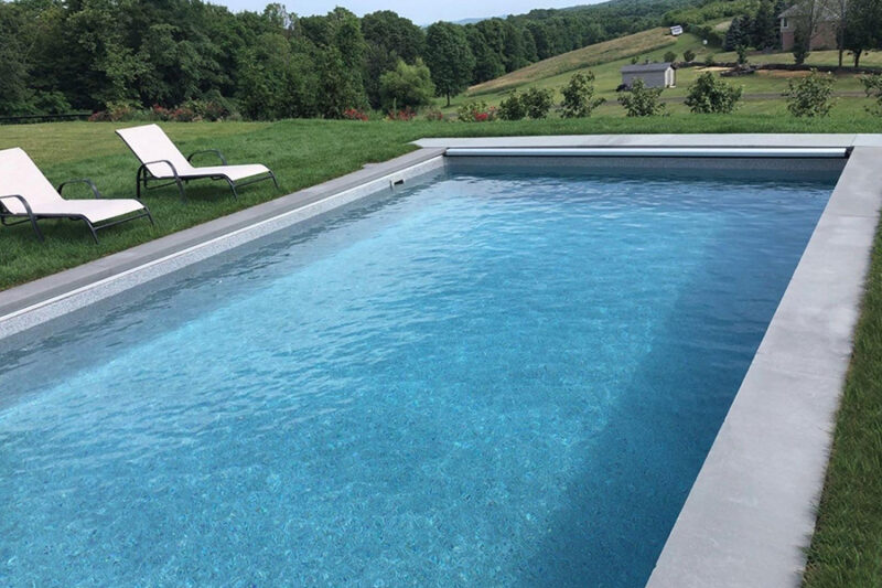 Rectangular inground fiberglass pool with clear blue water, surrounded by lush greenery and lounge chairs in a serene backyard