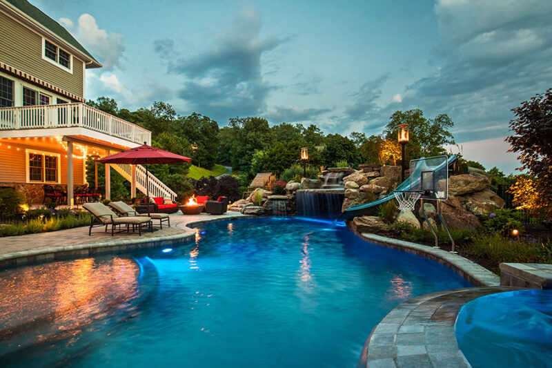 Luxurious backyard pool at dusk with slide, basketball hoop, and waterfalls, perfect for relaxation and entertainment