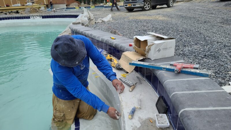 Worker performing gunite pool maintenance, applying material at pool's edge with tools nearby