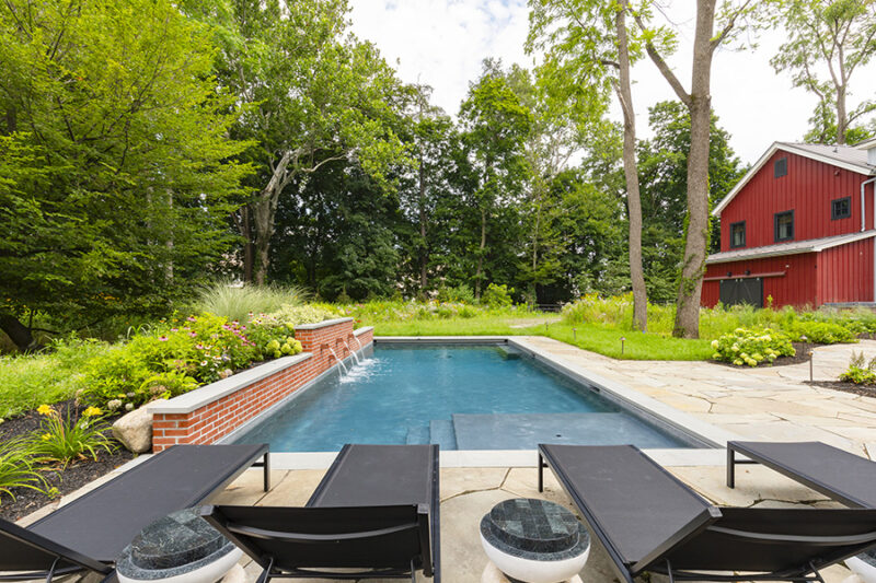 Backyard with a rectangular fiberglass pool shape, stone patio, lounge chairs, and a rustic red barn in the background