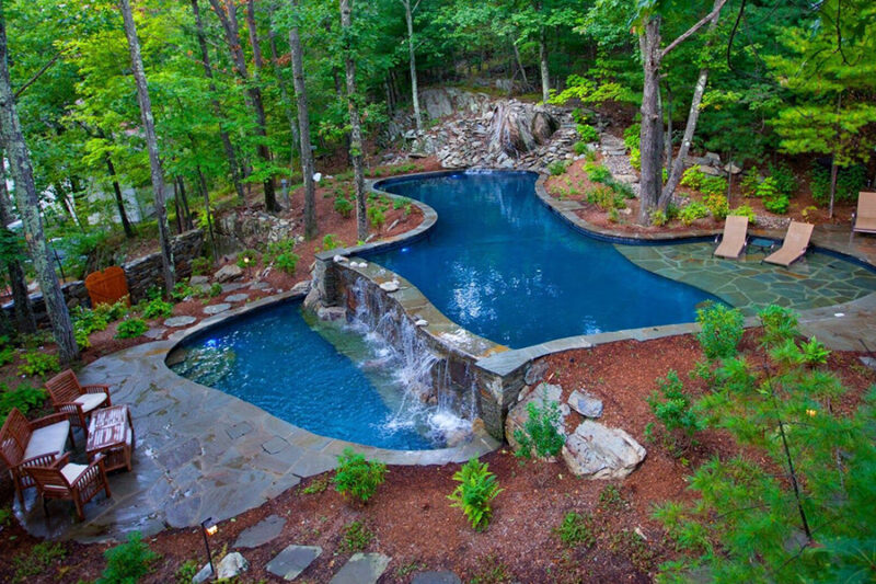 Gunite pool in a wooded backyard with a waterfall, stone patio, and seating area, showcasing a natural, luxurious outdoor space.