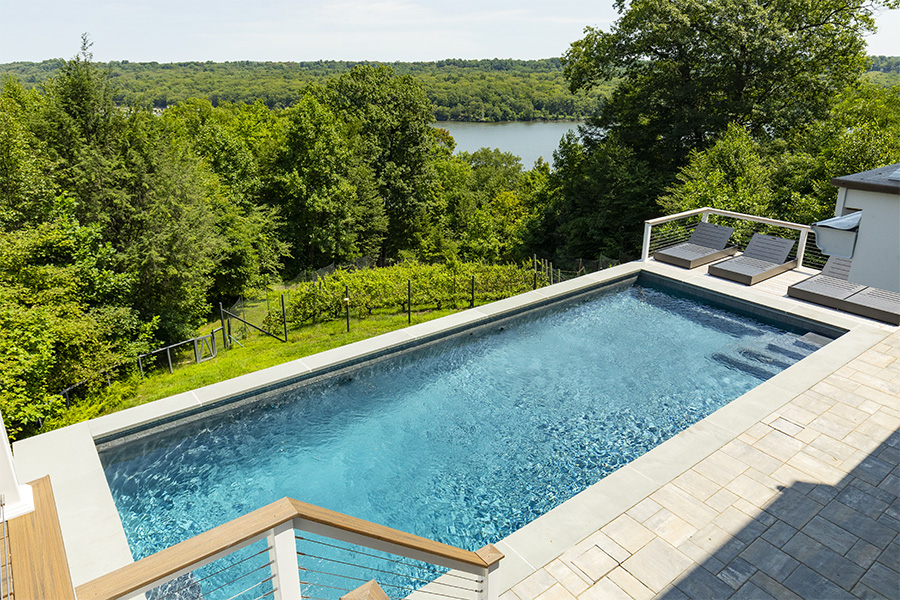 Modern rectangular pool design with light stone decking, lounge chairs, and a scenic lake view surrounded by lush greenery.